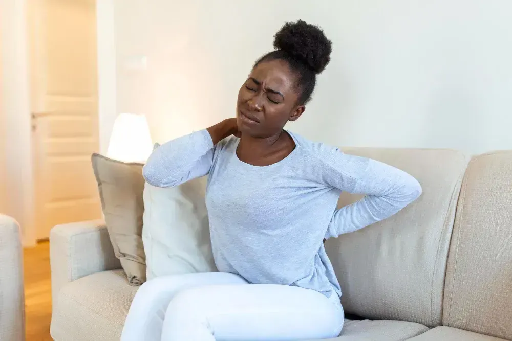 Mulher está no sofá e sente dores de fibromialgia pelo corpo.