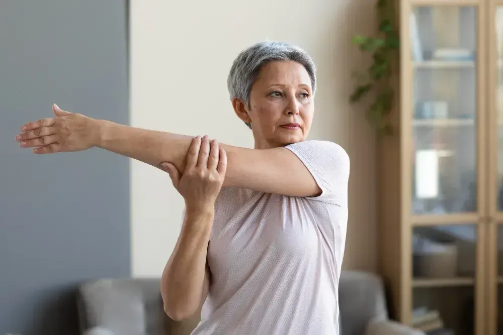 Mulher madura alonga o braço em casa em exercício relacionado à sarcopenia.